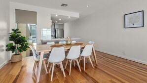 A modern dining area with a wooden table and white chairs, a potted plant, and a kitchen in the background. - Sea Star - Aldinga Beach - C21 SouthCoast Holidays (Aldinga Beach)
