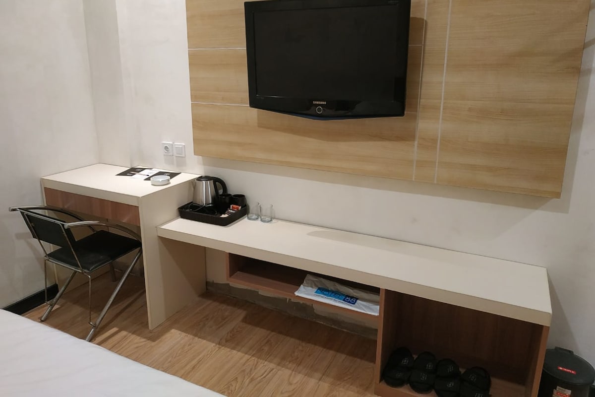 Habitación de hotel compacta con televisión de pantalla plana en la pared, un pequeño escritorio con silla, cafetera y zapatero.