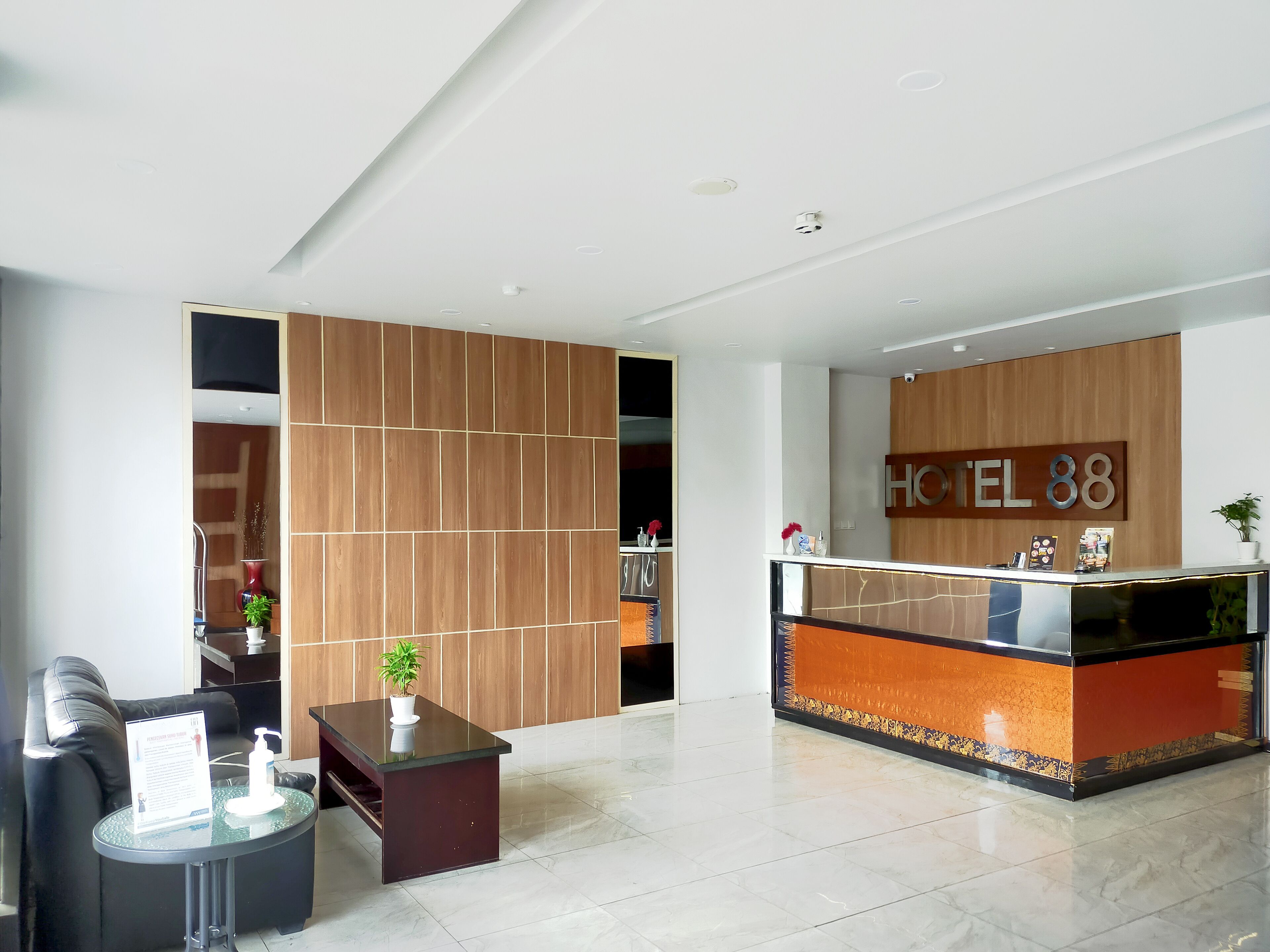 Zona de recepción de hotel con diseño moderno, que incluye un mostrador de recepción, un área de estar con sofá y una mesita, y un muro divisorio de madera.