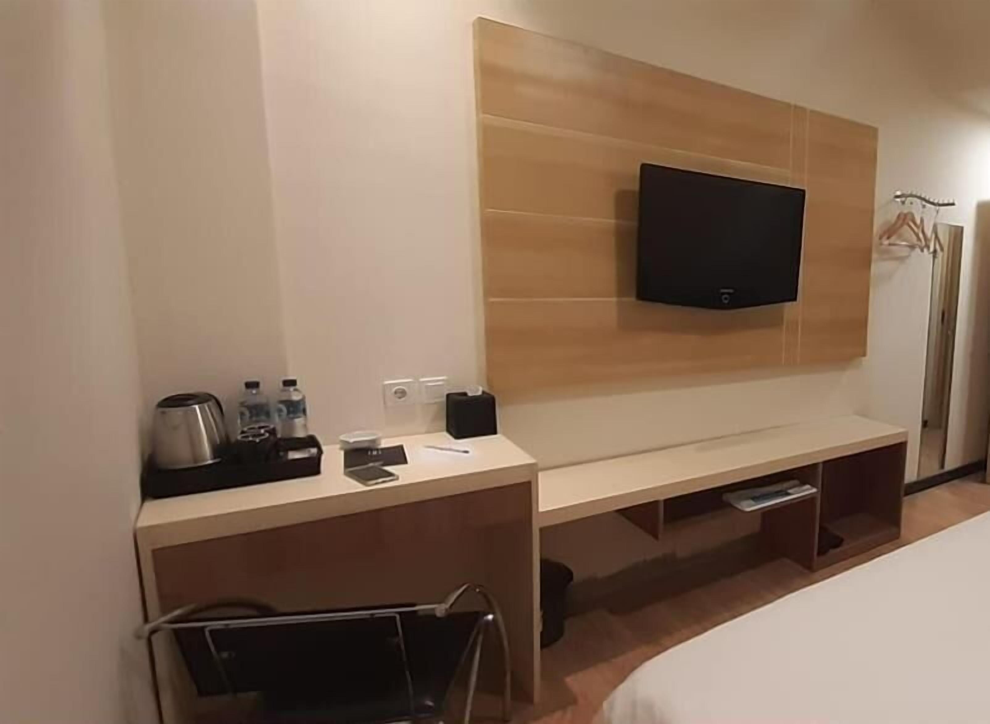 A hotel room with a flat-screen TV mounted on the wall, a bedside table with a lamp, a small desk with a chair, and a bed.