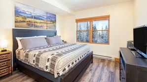 A bedroom with a large bed, a TV, a window with a view of trees, and a painting on the wall.