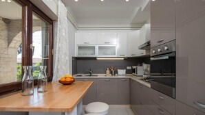 Una cocina compacta con encimera de madera, dos taburetes, un ventanal con cortinas de encaje y electrodomésticos modernos.