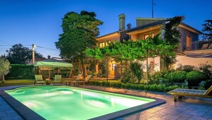 Una piscina con un borde de baldosas, rodeada de un jardín cuidado y una casa con balcón.