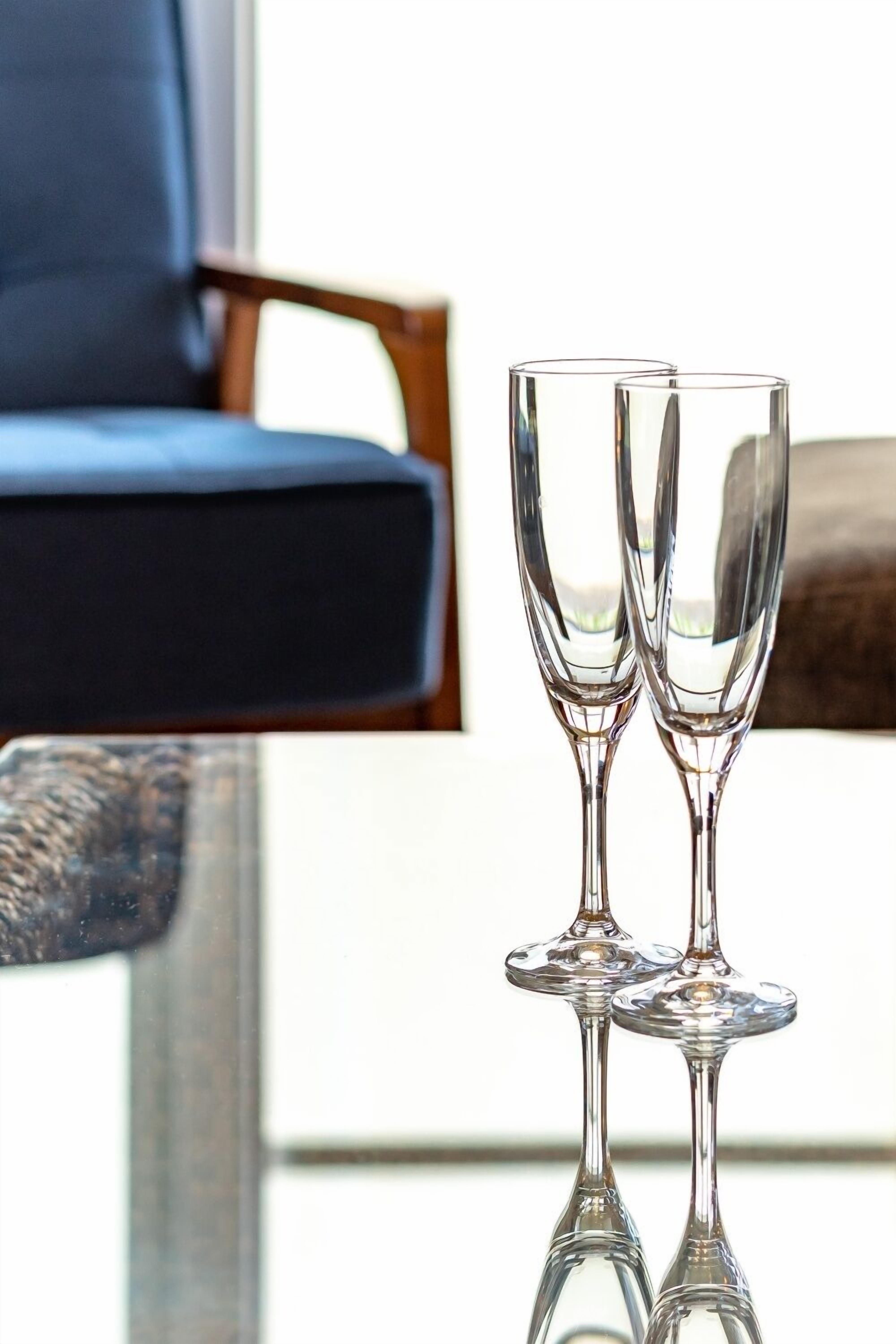 Deux verres transparents posés sur une surface réfléchissante, une chaise bleue et un accoudoir en bois.