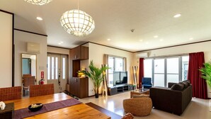 A modern living room with a wooden dining table, comfortable seating, a television, and large windows with curtains. - LiB PLUS Villa NAGO (Nago)