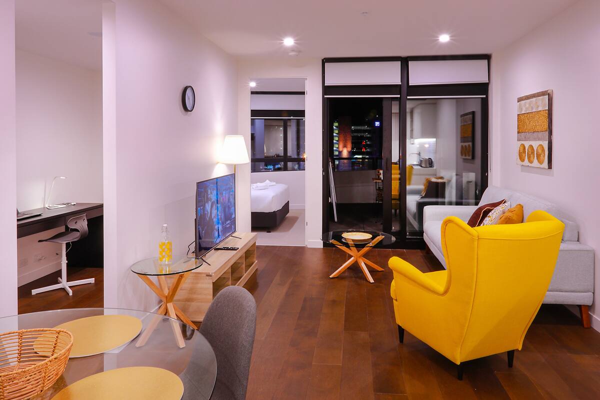 A modern hotel room with a bed, a yellow armchair, a glass table, and a desk with a computer.