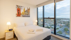 Ein Hotelzimmer mit einem großen Fenster, einem Bett, einem Nachttisch mit Lampe und einem Gemälde einer roten Telefonzelle.