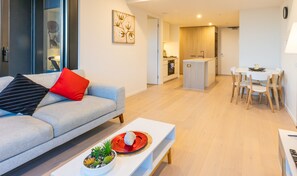A modern living room with a grey sofa, a red pillow, and a white coffee table with a plant. There is a dining area with a white table and chairs, and a kitchen in the background. - Astra Apartments Glen Waverley @ O'Sullivan RD (Glen Waverley)