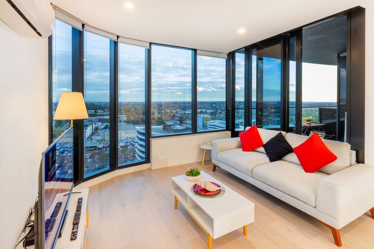 A modern living room with a large window, a white sofa with red and black cushions, a small white coffee table, and a lamp.