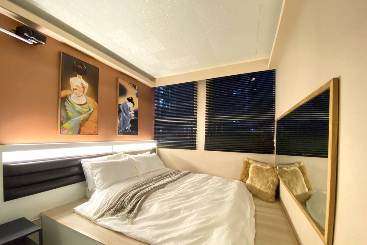 Une chambre à coucher moderne avec un lit, une table de chevet, une grande fenêtre avec des stores et des œuvres d’art murales.
