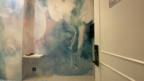 A room with a large abstract mural on the wall, a bed with a patterned bedspread, and a small shelf. - Guest House Conext (Kowloon)