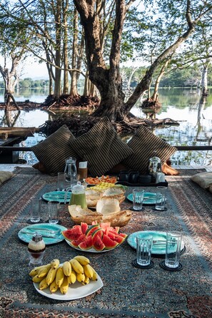 A table set outdoors with food and drinks, surrounded by trees and a body of water.