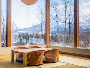 A dining area with a wooden table and wicker chairs, large windows with a view of snow-covered mountains and trees. - Aoyama Lodge (Kutchan)