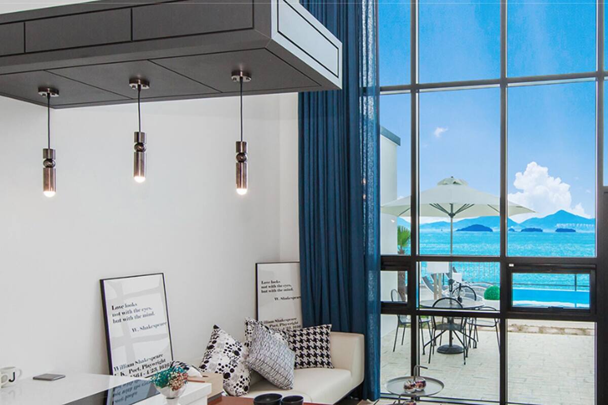 A modern room with a large window overlooking a beach, a ceiling light fixture, and a framed quote on the wall.