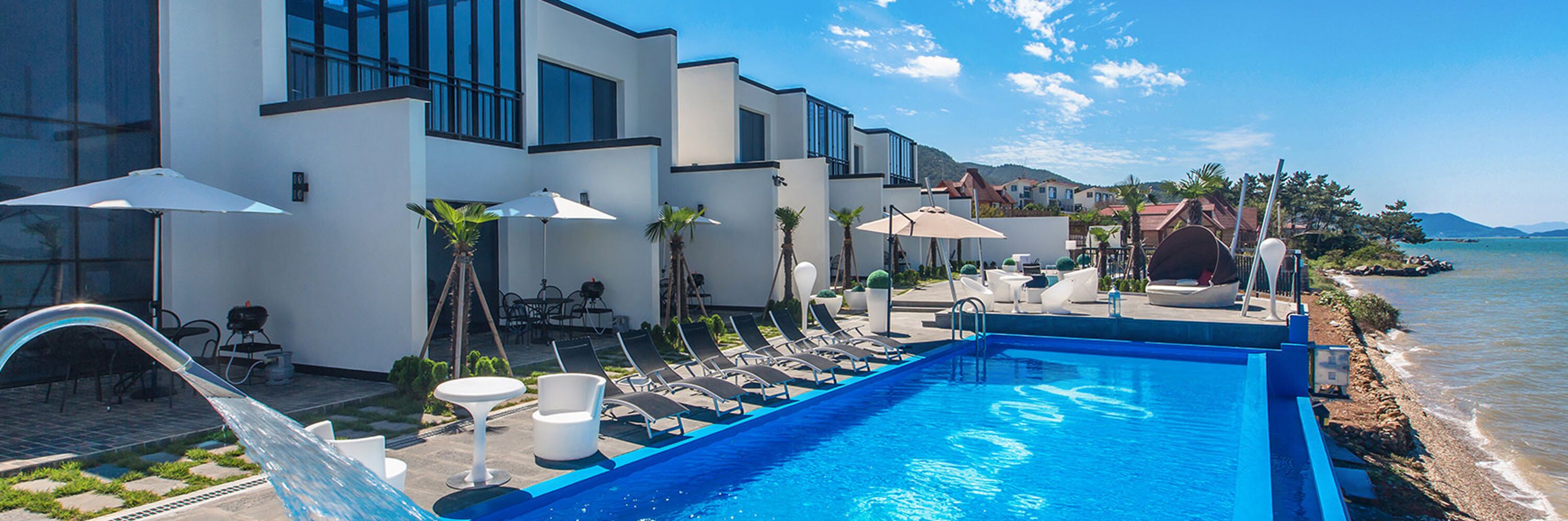 A modern hotel with a pool, sun loungers, umbrellas, and a view of the sea.