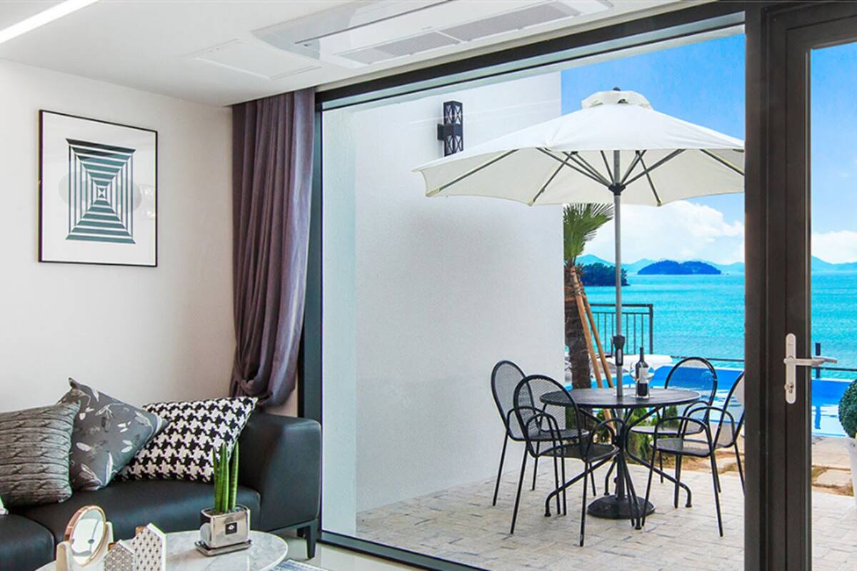 A balcony with a table, chairs, and an umbrella, overlooking a turquoise sea and distant islands.