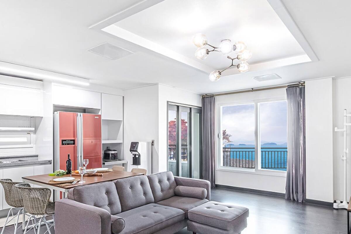 A modern apartment with a kitchen, dining area, and living room. It features a large window with a view, a gray sofa, and a dining table with chairs.