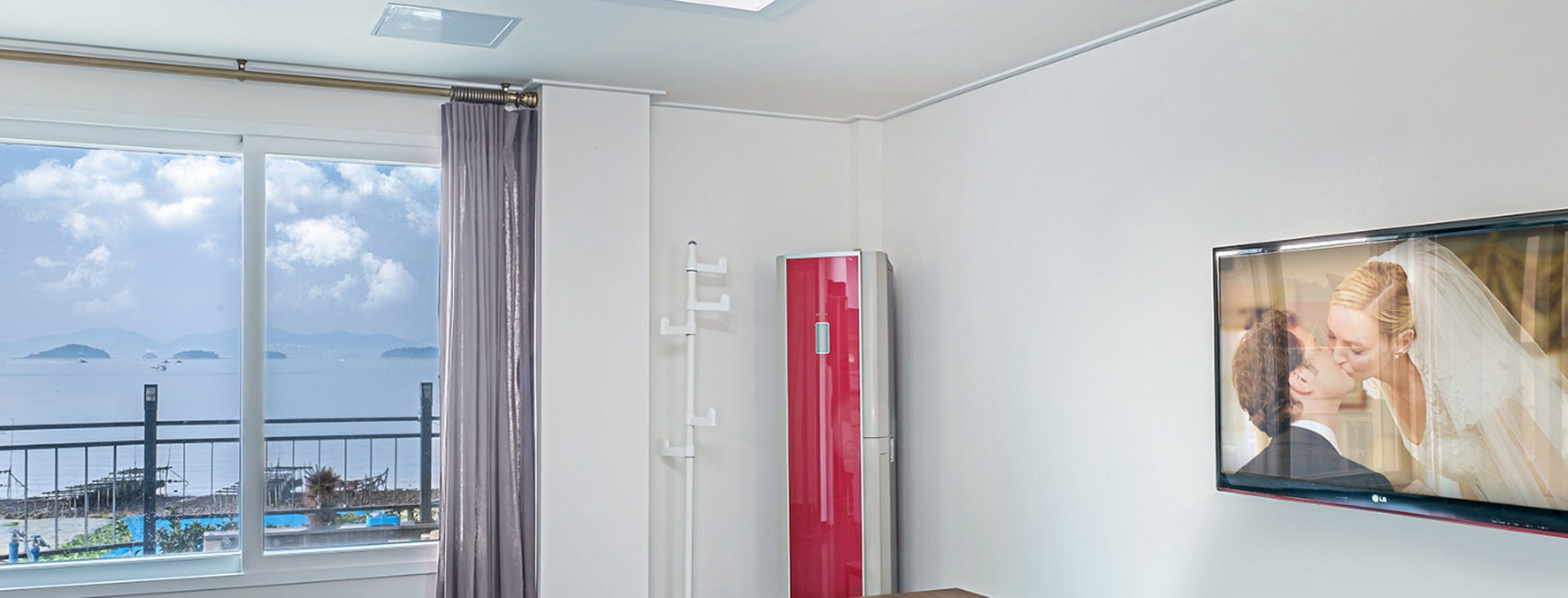 A room with a large window overlooking a body of water, a red air conditioning unit, and a framed photograph on the wall.
