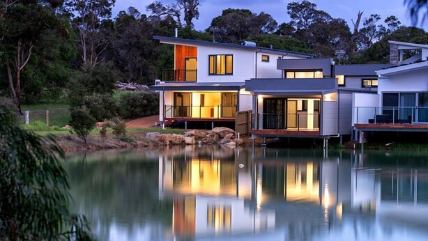 A modern house with a large deck overlooking a calm lake, surrounded by trees.