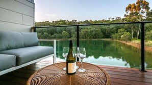 A balcony with a view of a lake, a bottle of wine, and two glasses on a woven table.