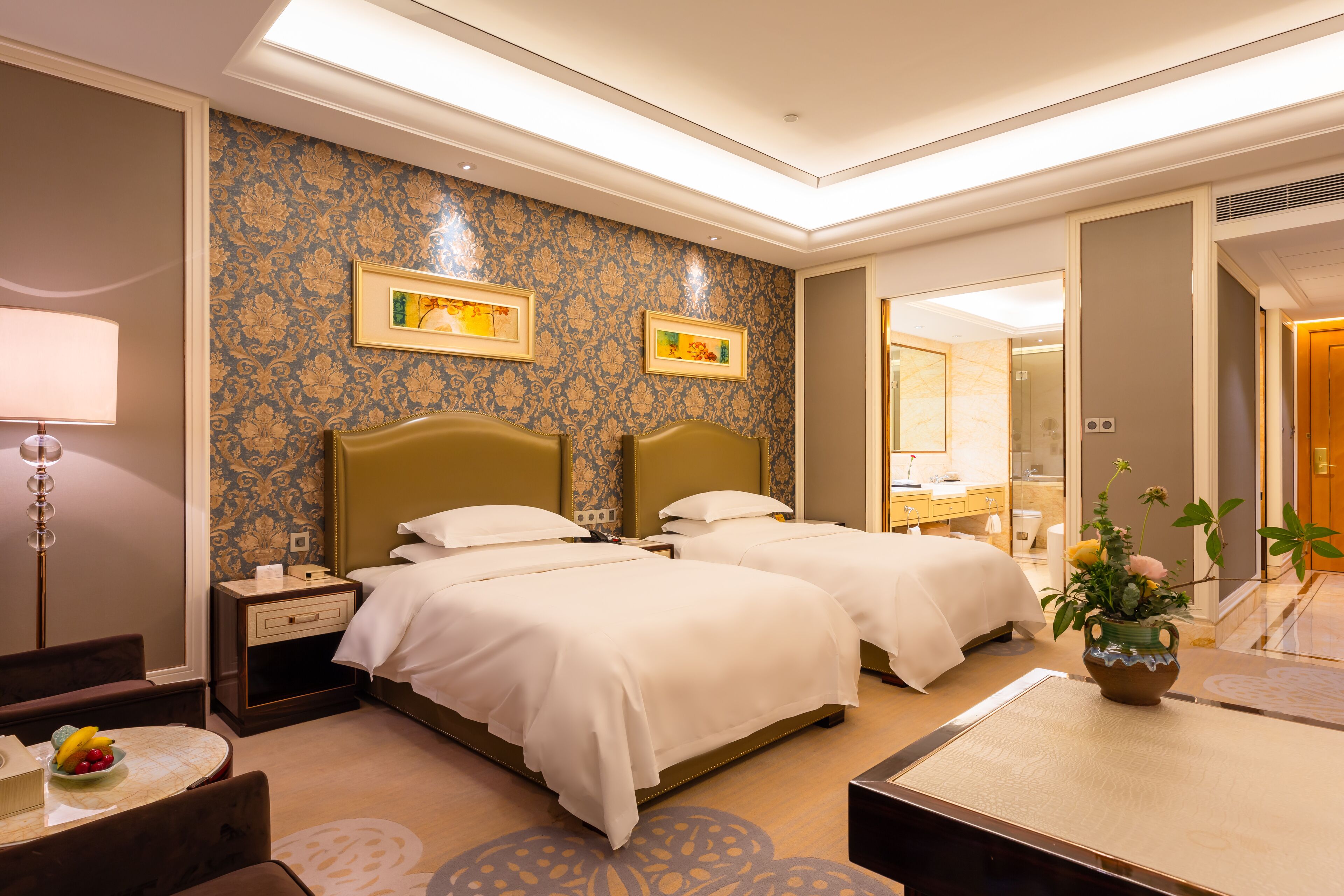 A hotel room with two beds, a nightstand, a desk with a lamp, and a vase with flowers.