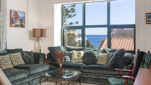 A living room with a large window, green sofas, a glass coffee table, and a painting on the wall.