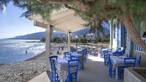 A seaside restaurant with outdoor seating, a covered patio, and a view of the sea and mountains.