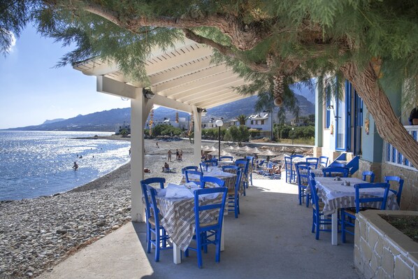 A seaside restaurant with outdoor seating, a covered patio, and a view of the sea and mountains.