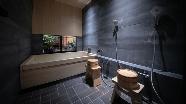 A modern bathroom with a wooden bathtub, a shower with a handheld showerhead, and a window with a view of greenery. - Tenyu (Kyoto)