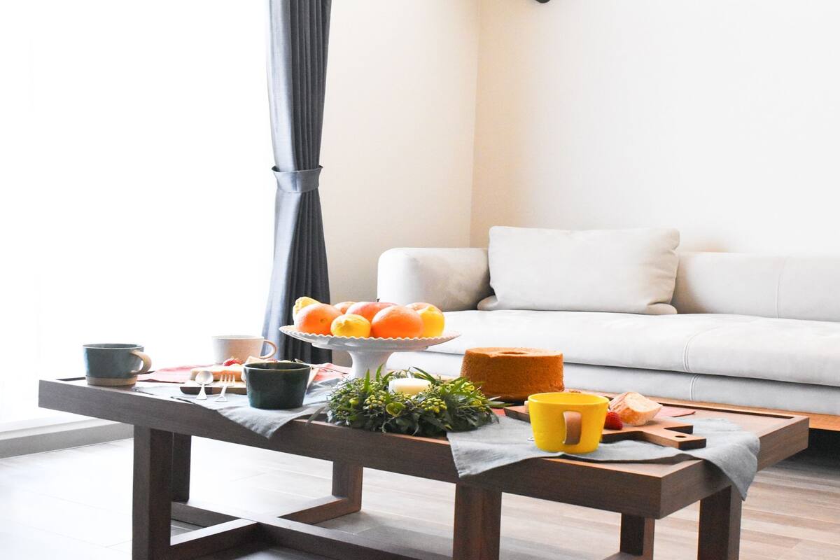 A modern living room with a white sofa, a wooden coffee table with breakfast items, and a window with curtains.