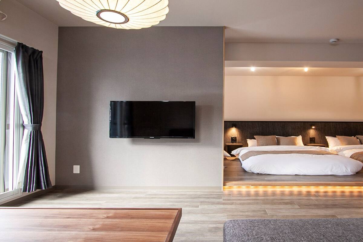A modern hotel room with a large bed, a flat-screen TV mounted on the wall, and a wooden floor.