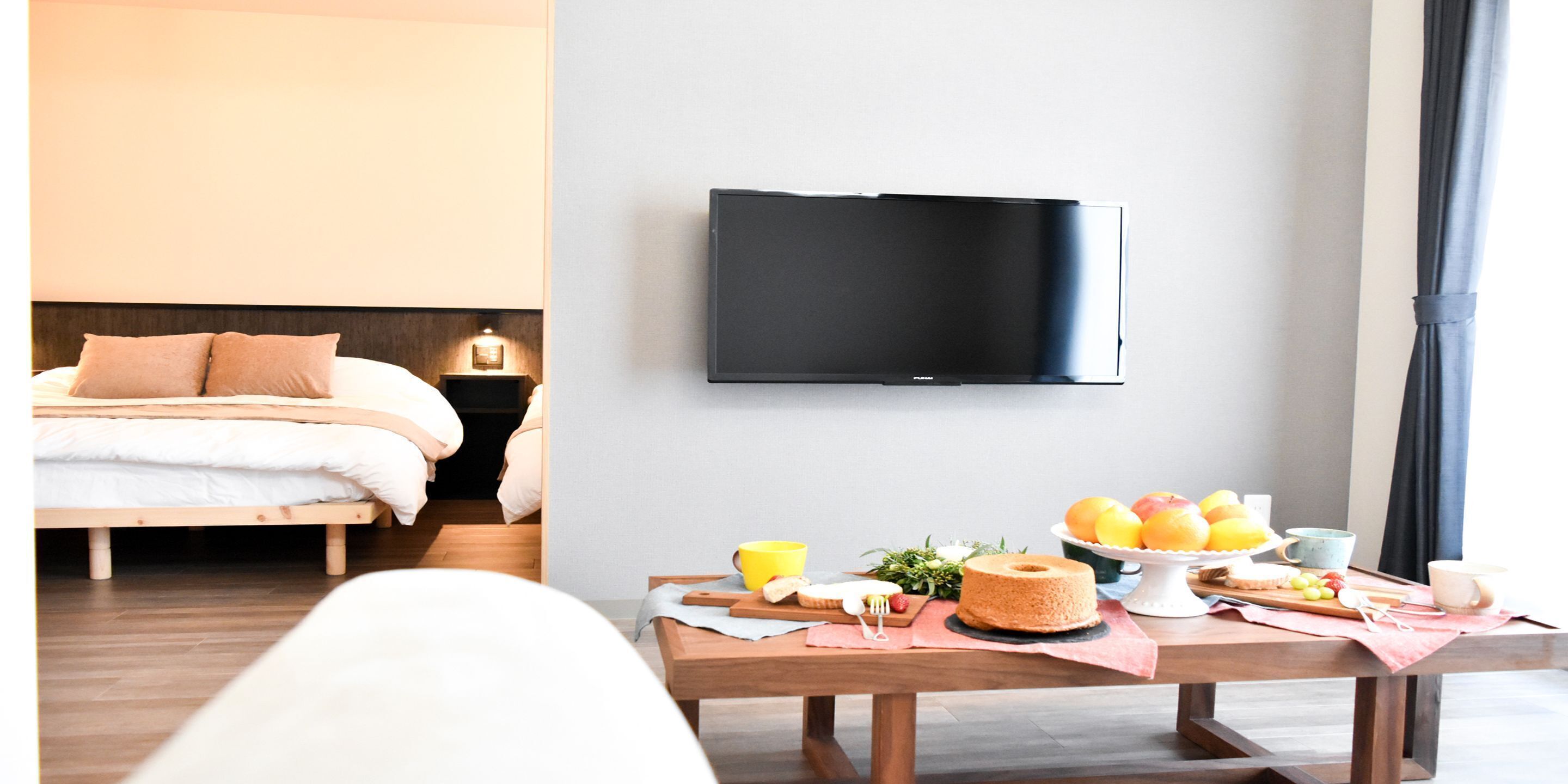 A hotel room with a bed, a bedside table with a lamp, a flat-screen TV mounted on the wall, and a breakfast table with fruits and a cake.