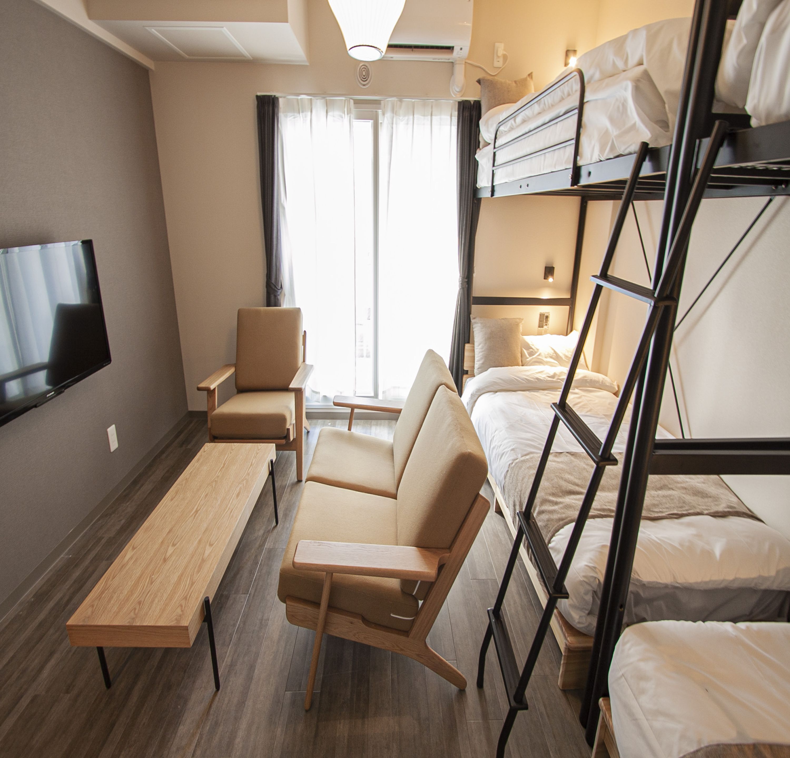 A hotel room with a bunk bed, a single bed, a desk, a chair, a TV, and a window with curtains.