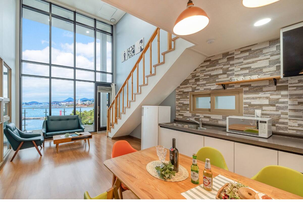 A modern kitchen with a stone backsplash, a dining area with a wooden table, and a view of the sea through large windows.