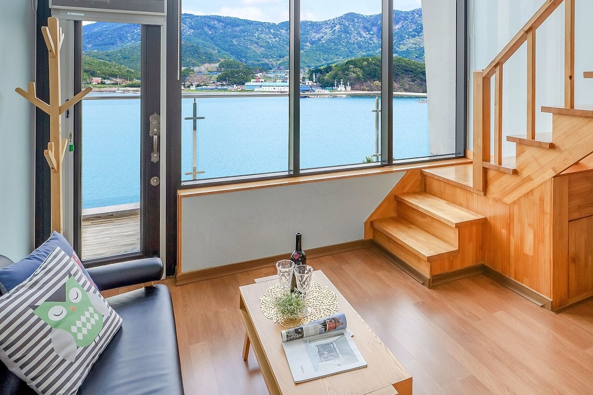 A modern living room with large windows, a wooden staircase, and a view of a lake and mountains.