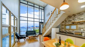 A spacious living area with a large window overlooking a body of water, a staircase leading to an upper level, and a kitchen area with modern appliances.