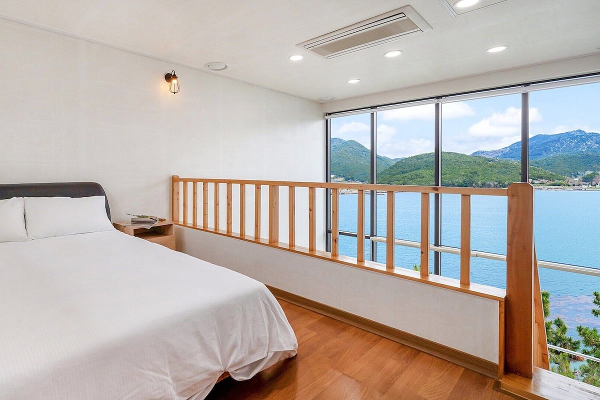 A bedroom with a large bed, a nightstand, a wooden railing, and a view of a lake and mountains.