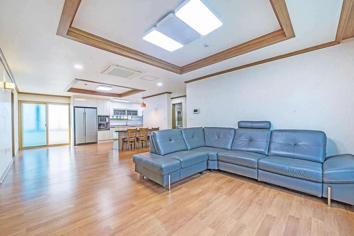 A spacious living area with a large L-shaped blue leather sofa, wooden flooring, and a modern kitchen in the background.