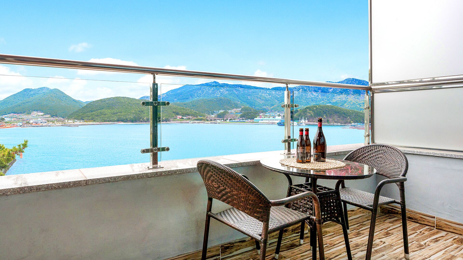 Ein Balkon mit Korbstühlen, einem Tisch und Flaschen, mit Blick auf eine Küstenlandschaft und Berge.