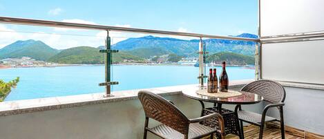 Ein Balkon mit Korbstühlen, einem Tisch und Flaschen, mit Blick auf eine Küstenlandschaft und Berge.