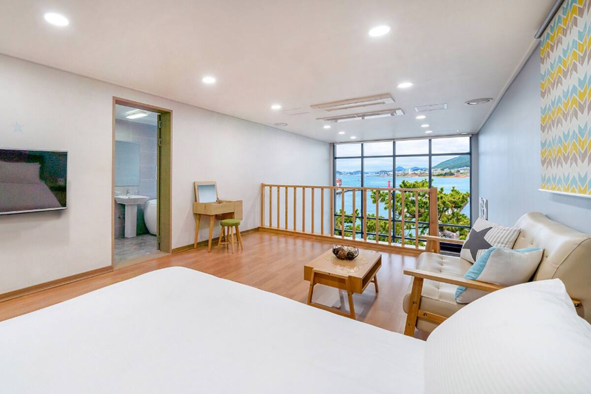 A spacious bedroom with a large bed, a TV mounted on the wall, a balcony with a view of the sea, and a small desk area.