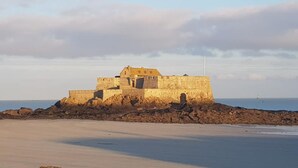 Uma fortificação costeira com uma torre central imponente e uma muralha ao redor, localizada numa praia arenosa com um mar calmo ao fundo.