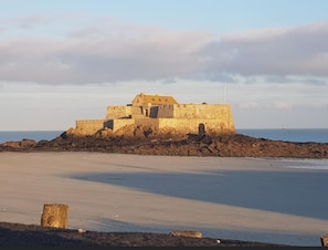 A coastal fortification with a prominent central tower and a surrounding wall, situated on a sandy beach with a calm sea in the background.