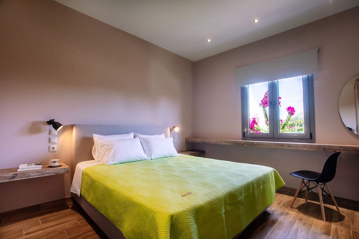 A bedroom with a bed, bedside table, lamp, chair, and a window with flowers.
