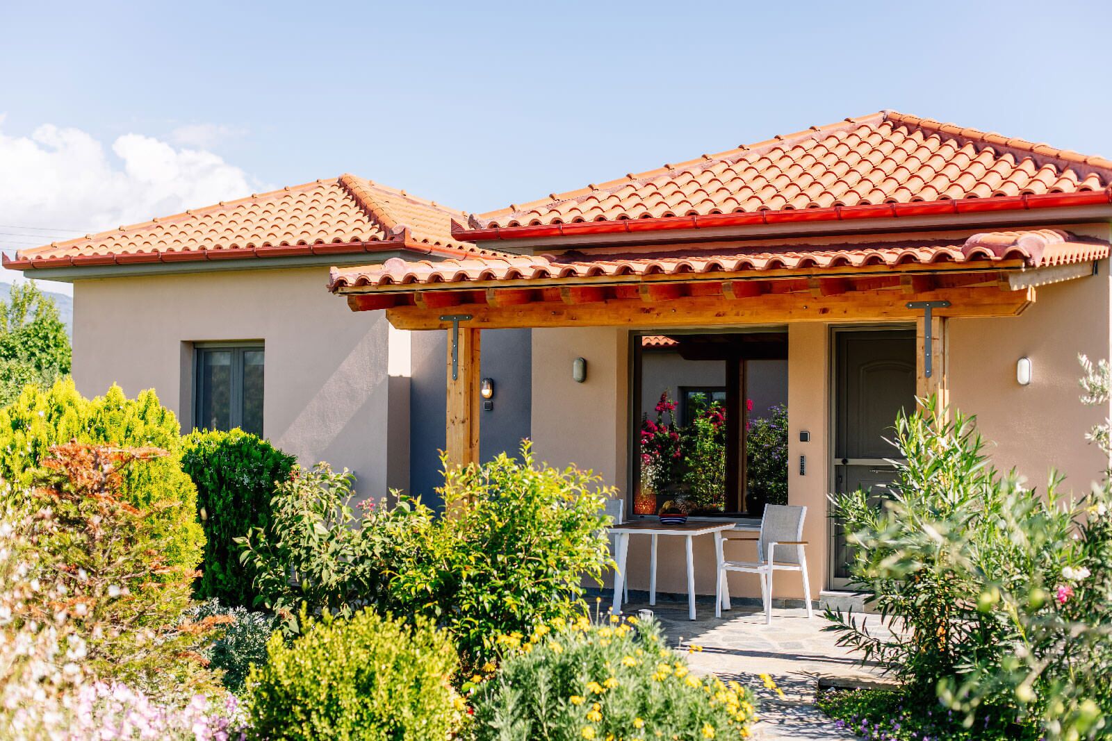 Une maison à deux étages, avec un toit en tuiles, un patio couvert doté d’une table et de chaises, et un jardin où poussent diverses plantes et fleurs.