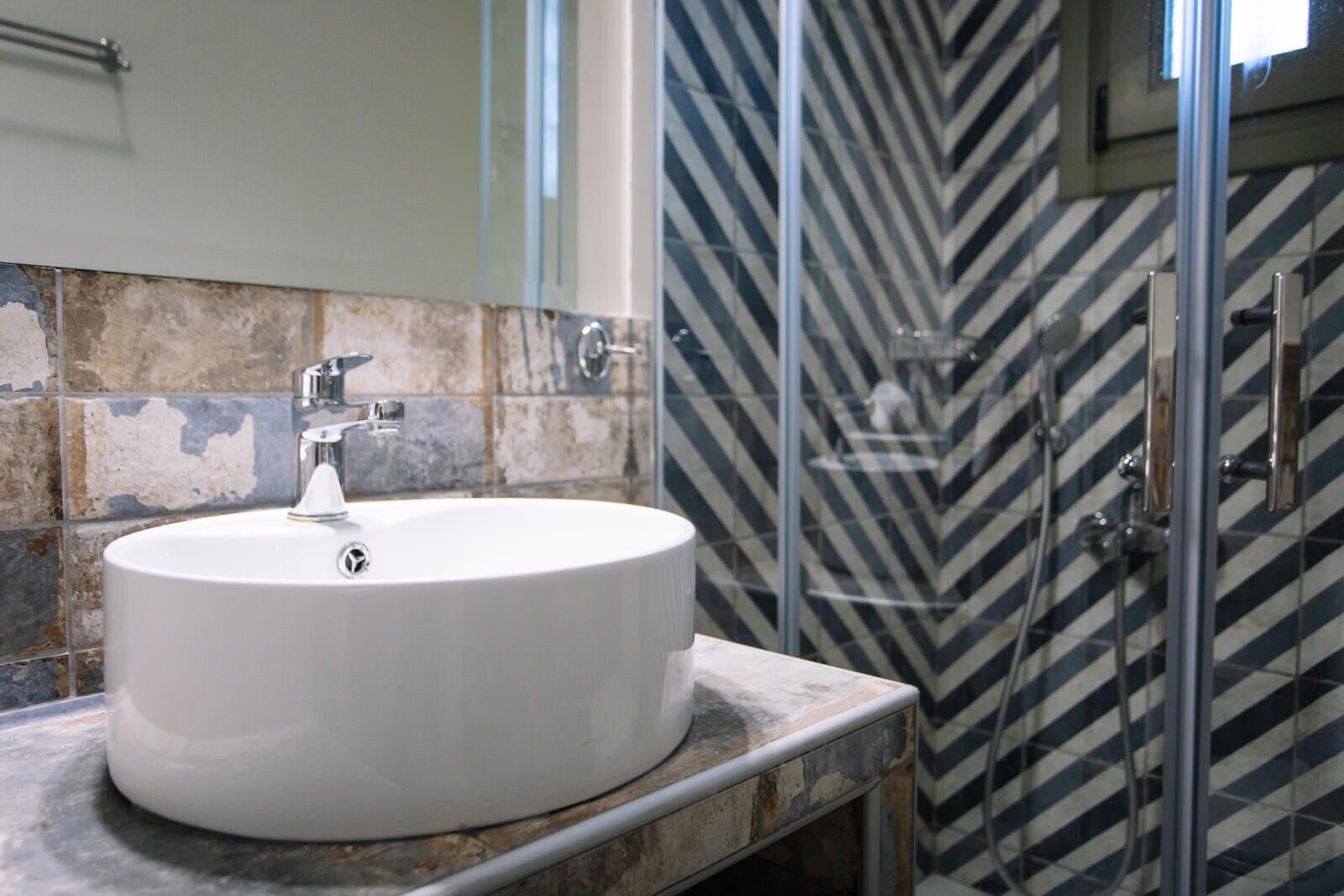 A bathroom with a round white sink, a shower with a glass door, and a tiled wall with a chevron pattern.