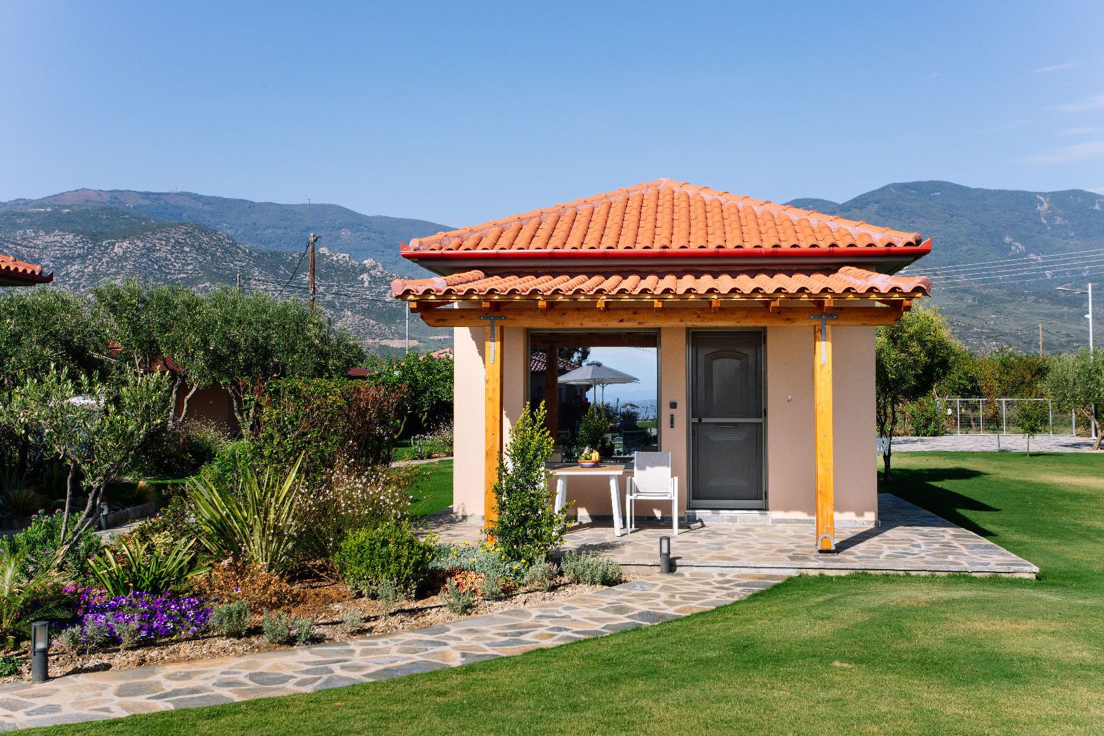 A covered outdoor seating area with a tiled roof, surrounded by a garden with various plants and a stone pathway leading to a door.