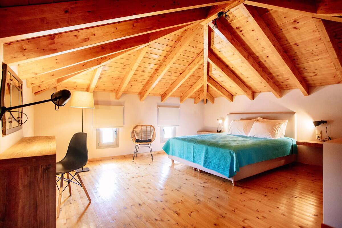 A bedroom with a sloped wooden ceiling, a bed with a teal bedspread, a desk with a lamp, and a chair.