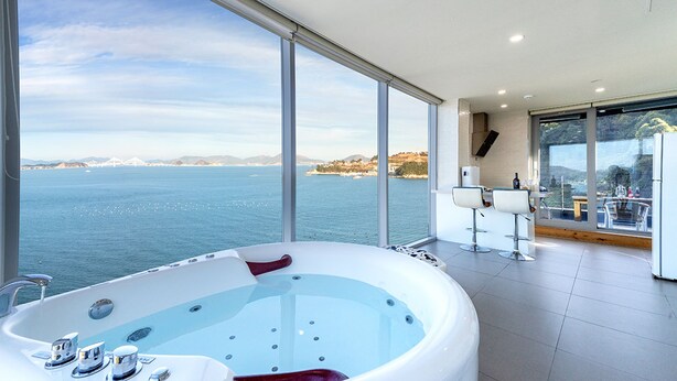 A modern bathroom with a large bathtub, a glass-enclosed balcony with seating, and a view of the sea and distant land. - House of Mind Pension (Geoje)