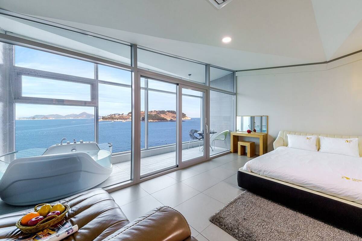 A modern bedroom with a large bed, a sitting area with a view of the sea, and a bathtub.